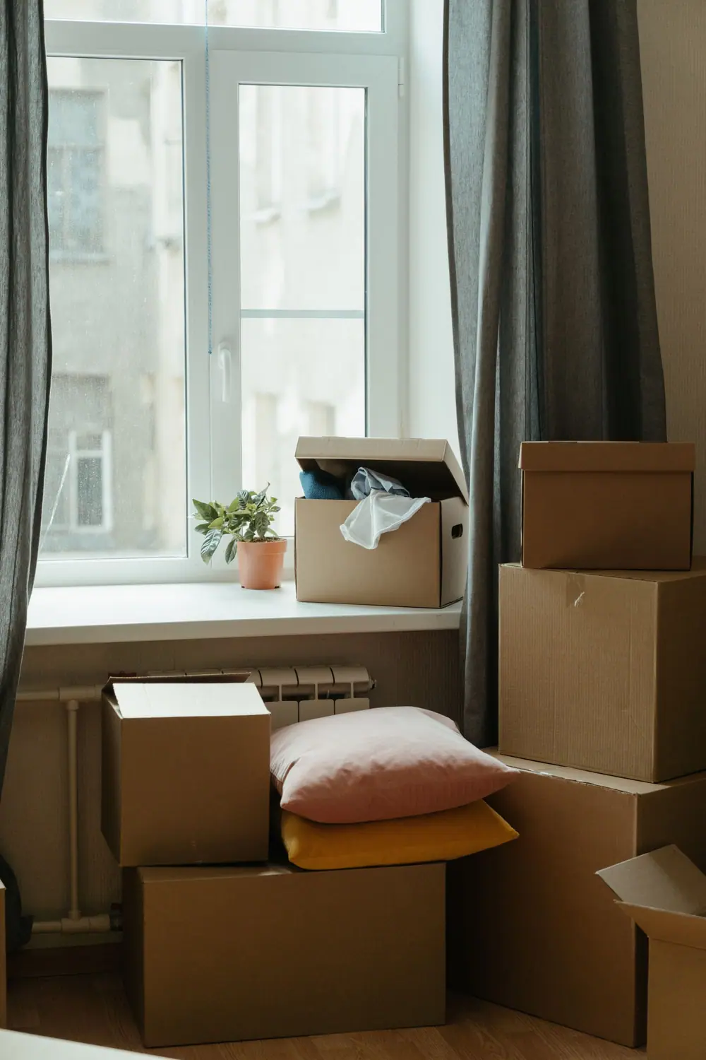 Stacked cardboard boxes and cushions beside a window and a radiator