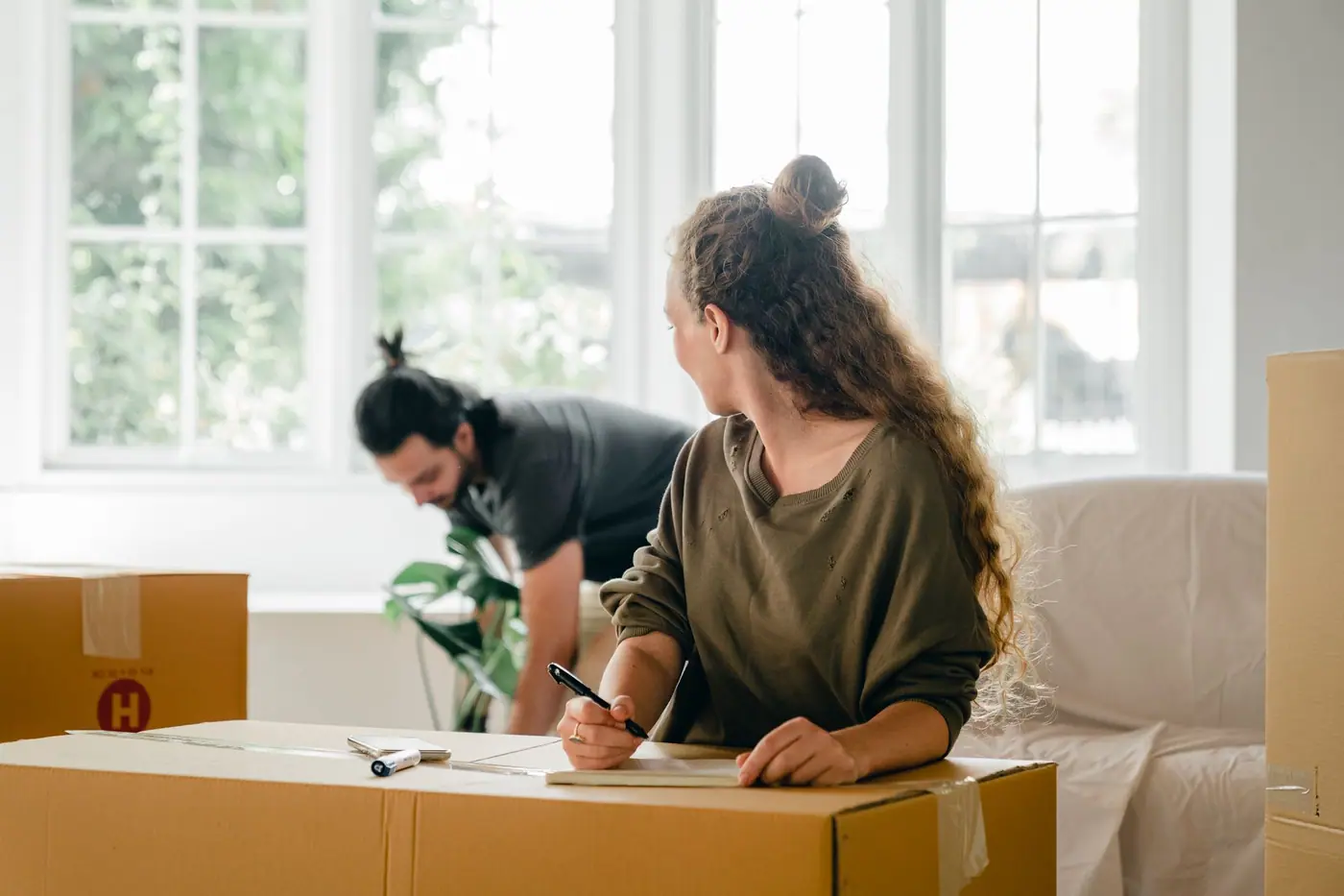 Two people with cardboard cartons in a bright room, one writing on a box
